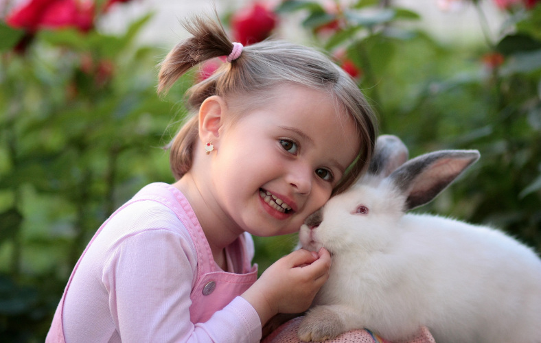 A smiling child gently nuzzles a white rabbit among roses